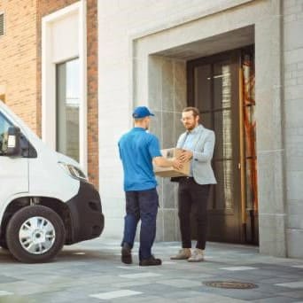 Kurier in blauer Arbeitskleidung reicht einem Mann einen Karton vor einem modernen Bürogebäude.