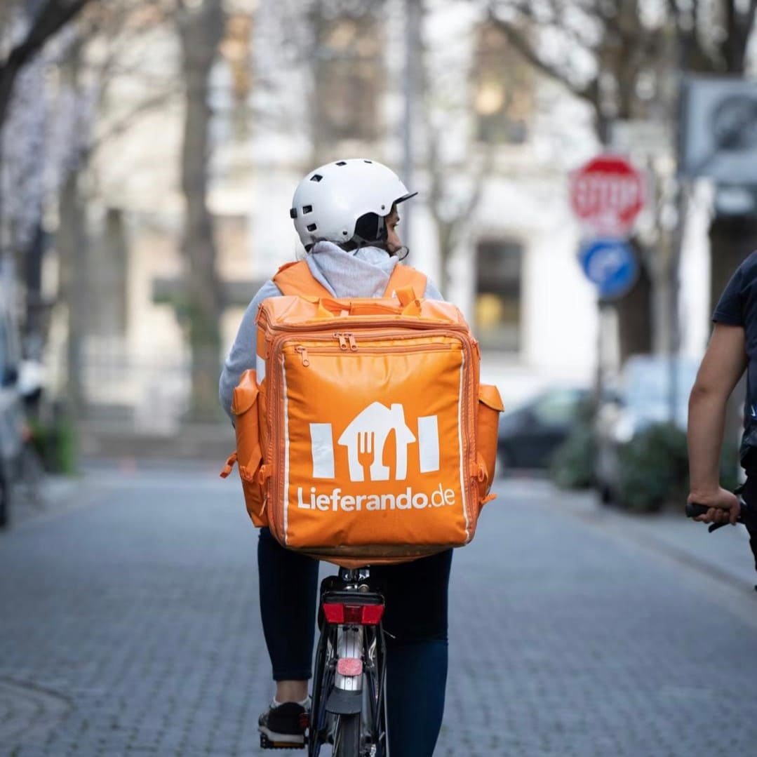 Fahrradkurier von Lieferando mit orangefarbenem Lieferrucksack auf einer Stadtstraße.